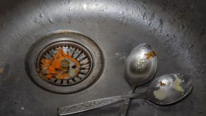 Close-up of a stainless steel sink with food scraps caught in the drain strainer and two dirty spoons with leftover grease and food residue nearby.