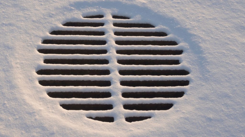 Round metal storm drain partially covered with snow, showing icy buildup around the grate during winter conditions.