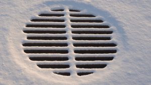 Round metal storm drain partially covered with snow, showing icy buildup around the grate during winter conditions.