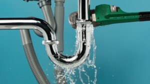 Close-up of a leaking chrome drain pipe being tightened with a green pipe wrench, with water spilling out against a blue background.
