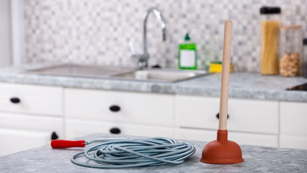 A drain snake and plunger placed on a kitchen countertop with a modern sink and backsplash in the background, representing DIY plumbing tools.