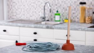 A drain snake and plunger placed on a kitchen countertop with a modern sink and backsplash in the background, representing DIY plumbing tools.