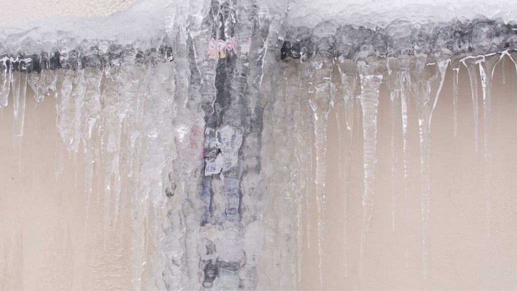 Winter Effects on Drain Pipes – Exterior drain pipes covered in thick ice and icicles, showing severe freezing during winter weather.