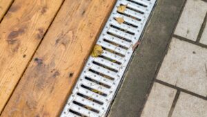 Close-up of a metal drainage grate surrounded by wooden decking and concrete pavers, with fallen leaves partially blocking the drain openings.
