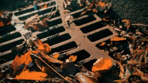 A close-up of a catch basin cover with fallen autumn leaves and debris scattered over it. The image shows the metal grid of the drain cover partially covered by dry leaves, indicating the need for cleaning and maintenance. The overall lighting suggests a late afternoon or early evening setting.