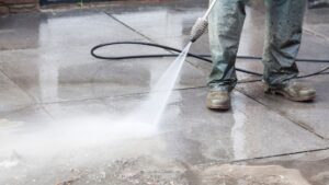 A worker in waterproof clothing and boots uses a high-pressure washer to clean a wet concrete surface, likely part of an underground garage or pavement area.