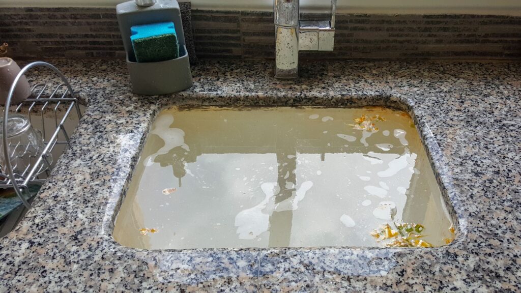 A clogged kitchen sink filled with dirty water, soap suds, and debris. The sink is made of granite with a faucet visible on the right side, and a dish rack and sponge holder are seen nearby.