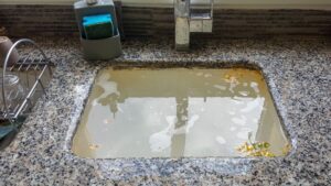 A clogged kitchen sink filled with dirty water, soap suds, and debris. The sink is made of granite with a faucet visible on the right side, and a dish rack and sponge holder are seen nearby.